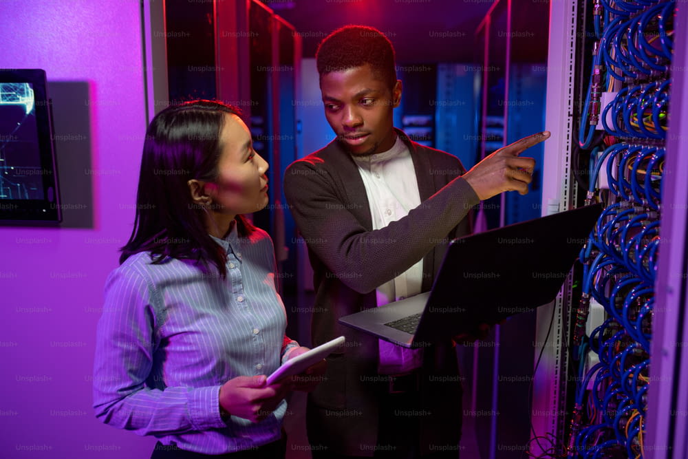 Young Afro-American technician pointing at cables while explaining server productivity to Asian colleague in data center illuminated with neon light
