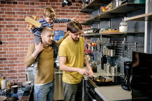 Giovane uomo gay che cucina la colazione sul fornello in cucina quando suo marito dà un giro in piggyback al loro piccolo figlio