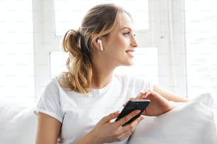 캐주얼 티셔츠를 입은 20대 백인 소녀의 초상화는 집에서 밝은 방의 소파에 앉아 휴대폰을 사용하여 휴대폰을 사용한다