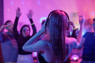 Vista trasera de una mujer joven con cabello largo y rubio tocando auriculares mientras está parada frente a una multitud de amigos emocionados bailando en la fiesta