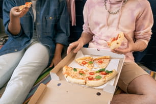 Two relaxed teenagers in casualwear eating pizza from box while sitting on the floor