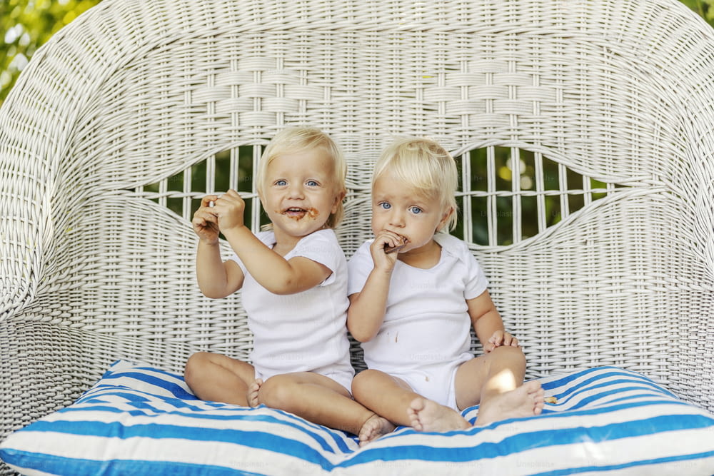 Toddler twins are on the wicker chair in the garden eating cookies. Cute babies in the white baby bodies hold a cake in their hands. Light hair, blue eyes, toddler babies, peaceful childhood