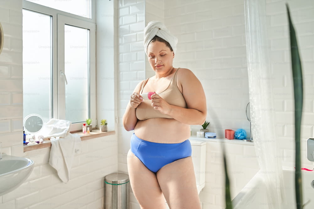 Vue pleine longueur de la femme portant de la lingerie regardant son antisudorifique debout à la salle de bain. Photo d’archives
