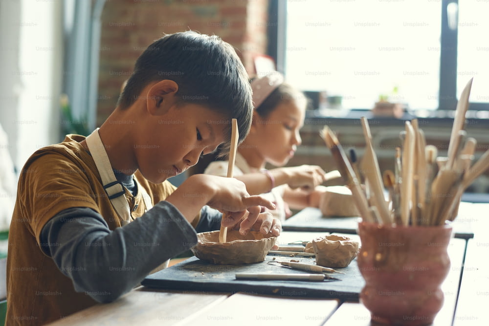 Ragazzo asiatico concentrato seduto a tavola e usando il bastone di legno mentre fa la ciotola di argilla alla lezione di ceramica