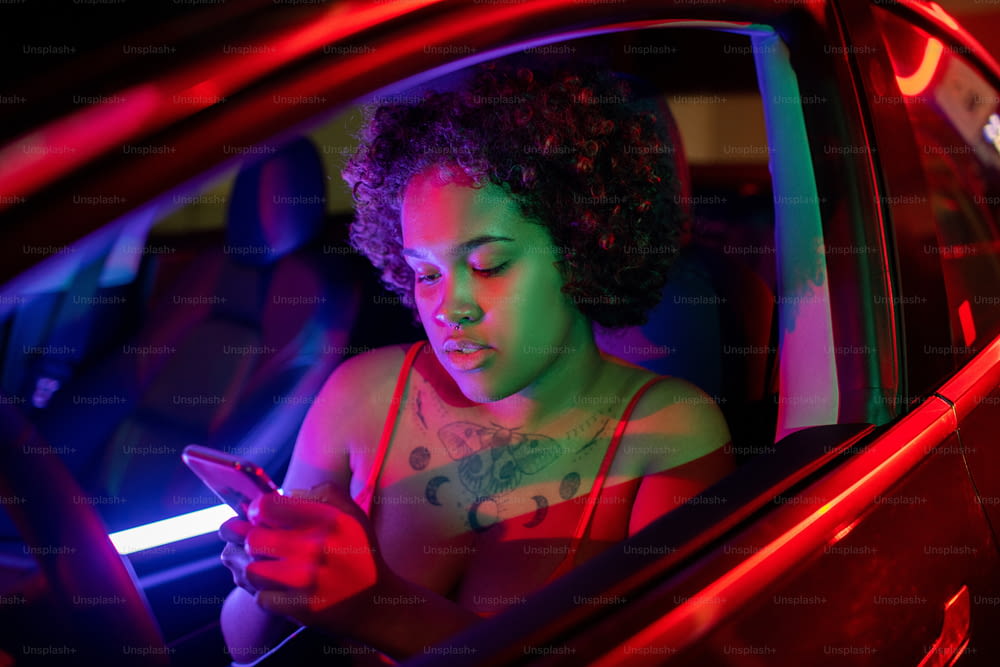 Menina africana bonita rolando no smartphone enquanto se senta na frente do volante no carro elétrico