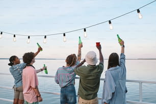 夏の日に水辺の前に立つ飲み物を持つ若い友達の背面図