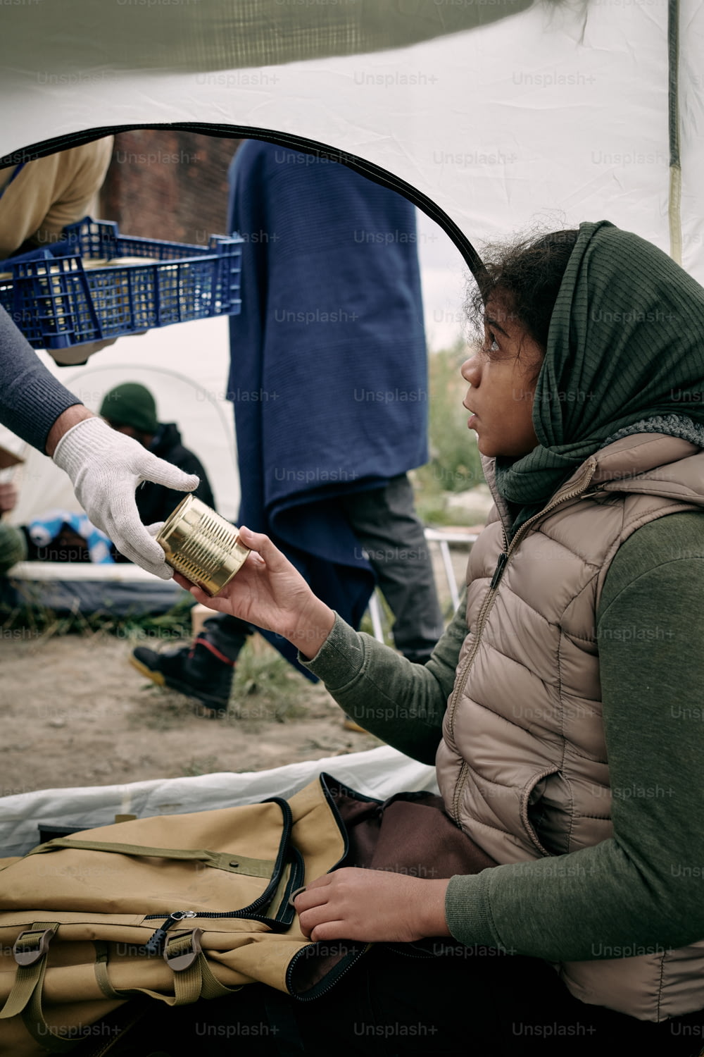 Une jeune réfugiée affamée prend de la nourriture en conserve de la main gantée d’un bénévole masculin assise dans une tente