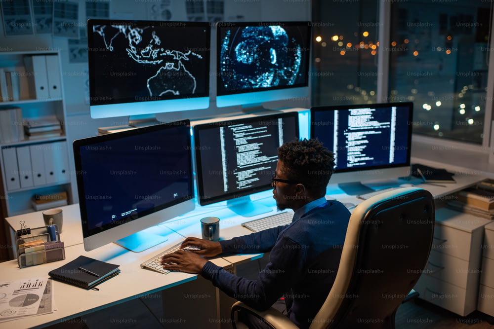 Giovane sviluppatore di software contemporaneo che lavora con dati decodificati mentre è seduto davanti al monitor del computer in ufficio di notte