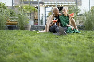 Multiracial couple eating watermelon in their garden. Concept of relationship and enjoying time together. Modern lifestyle. Black man hugging his european girlfriend. People covered in plaid