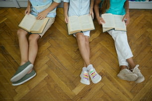 Favorite activity. Top view of legs of schoolchildren sitting on parquet floor with open books reading, no face
