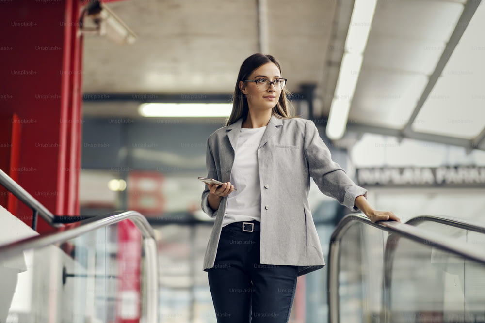 A young, elegant businesswoman descending the escalator and going into the underground garage. She is holding a phone and looking away. A workday of a businesswoman