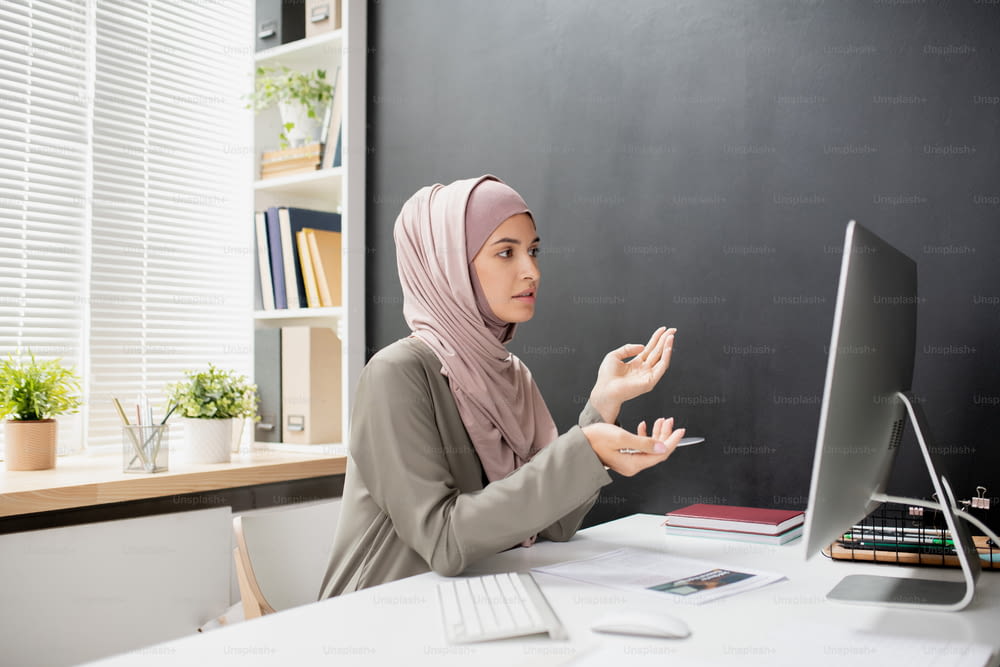 Young Muslim female teacher with pen explaining something to online audience while sitting in front of computer monitor