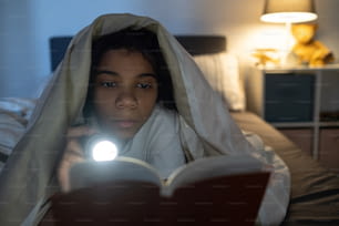 Chica afroamericana seria acostada debajo del edredón en la cama y linterna encendida en el libro en la oscuridad