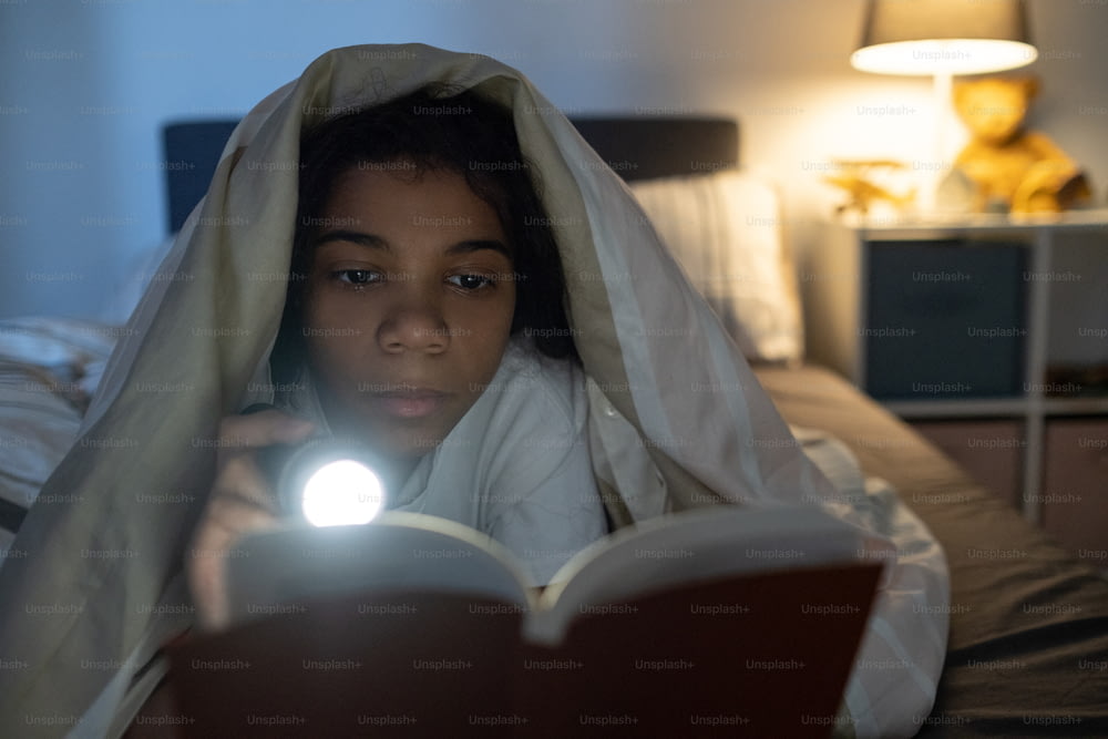 Serious African American girl lying under duvet on bed and shining flashlight on book in darkness