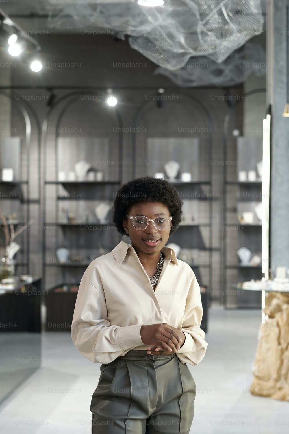 Happy young elegant shop assistant of jewelry boutique standing in front of camera on background of large display
