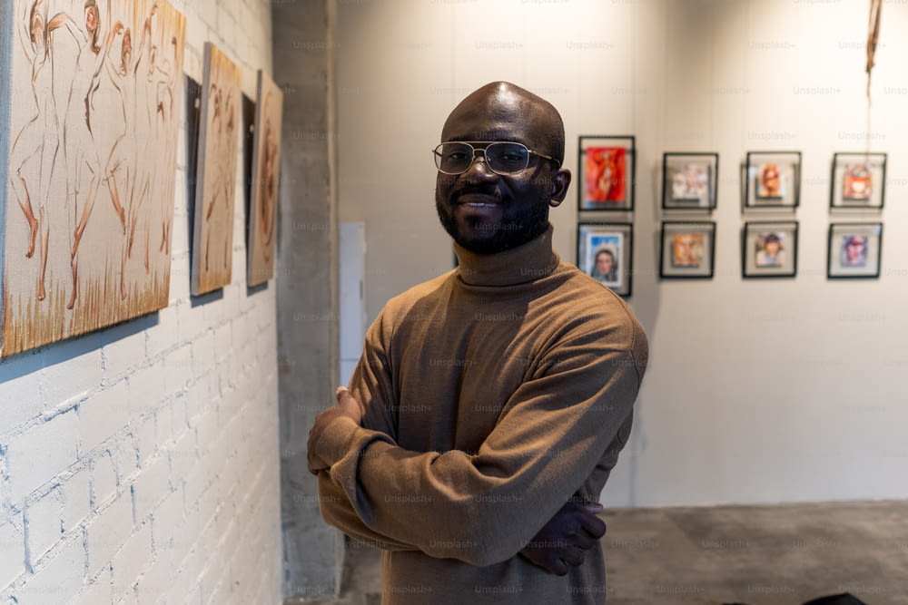 Portrait d’un jeune visiteur africain souriant à la caméra alors qu’il se tenait debout, les bras croisés près du mur avec des œuvres d’art dans un musée d’art