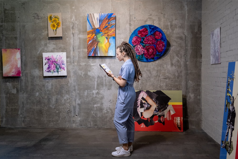 Jeune femme lisant un livret debout dans une galerie d’art avec de belles peintures sur le mur