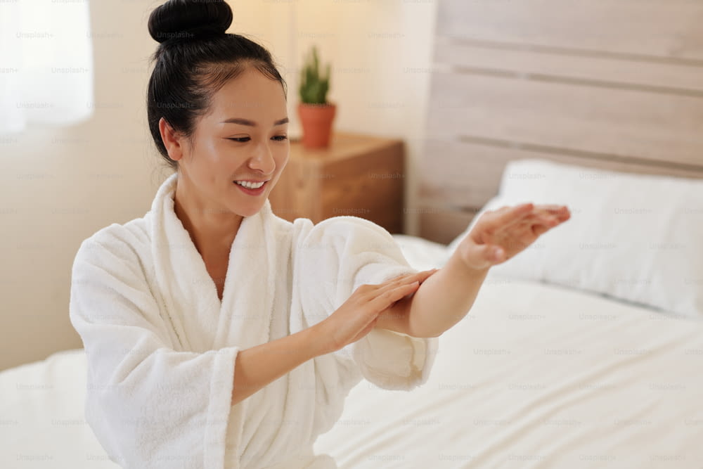 Happly giovane donna asiatica che applica olio per il corpo sulle braccia dopo aver fatto il bagno a casa la sera