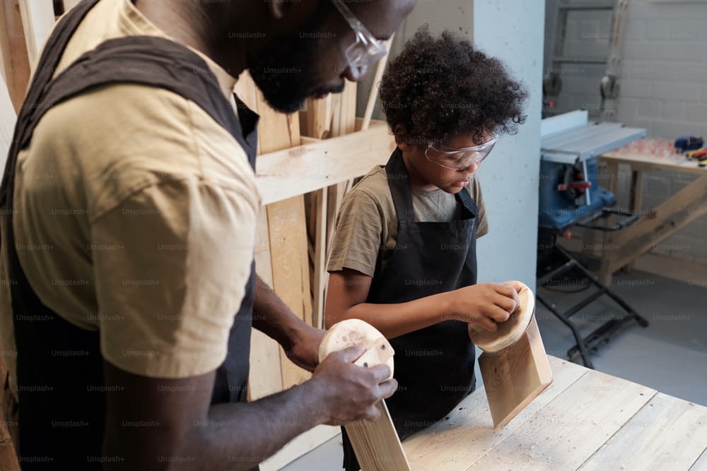 Portrait of black father and son sanding wood in carpentry workshop together, copy space