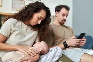 朝、スマートフォンで夫に対してベッドに座りながら、手に眠っている赤ちゃんを見ている若い美しい女性