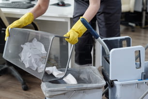 Behandschuhte Hände einer Putzfrau, die Müll aus dem Mülleimer in einen Plastikeimer auf dem Hausmeisterwagen wirft, während sie in einem modernen Büro arbeitet
