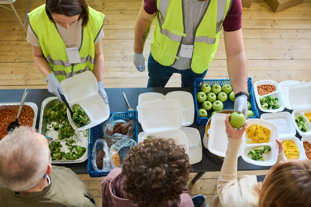 Sobre el ángulo de dos voluntarios uniformados repartiendo comida entre los refugiados con contenedores de pie en cola por mesa