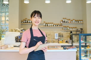 Portrait of Asian young waitress in apron smiling at camera and making notes in notepad while standing in coffee shop