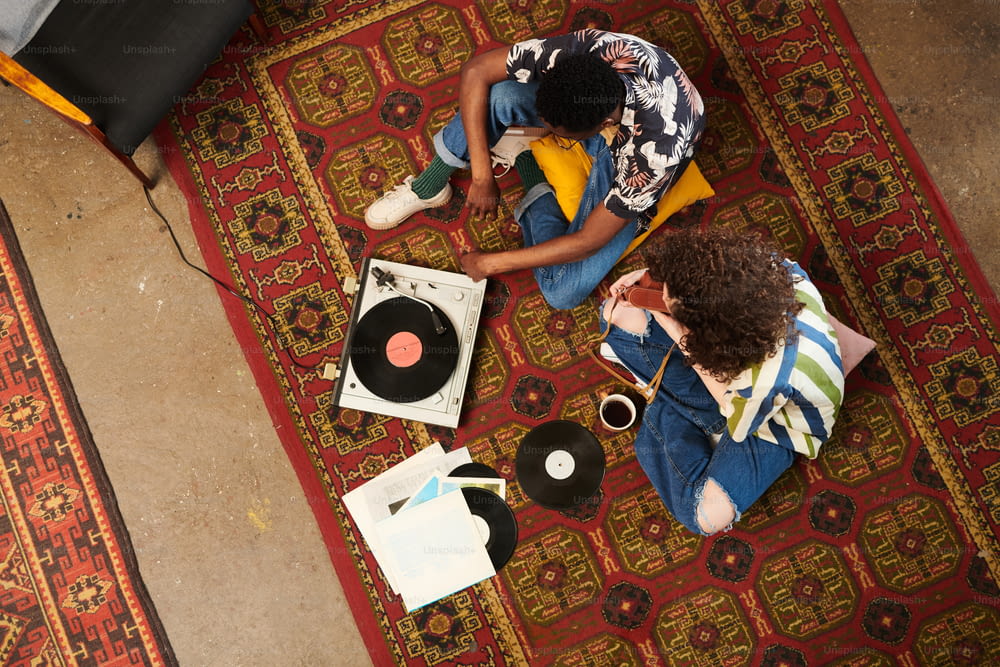 Über dem Winkel von zwei Zoomern in stilvoller Freizeitkleidung, die Vynil-Scheiben auf dem Plattenspieler hören, während sie sich auf dem roten Teppich im Wohnzimmer entspannen