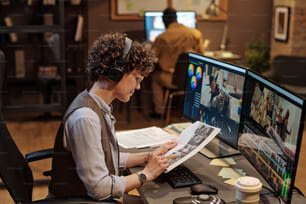 Female editor in headphones examining script at her workplace while editing photos on computer