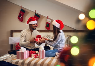 Glückliches, aufgeregtes, hübsches junges Paar, das sich gegenseitig Weihnachtsgeschenke macht, während es in Pullovern und Weihnachtsmützen auf dem Bett sitzt.