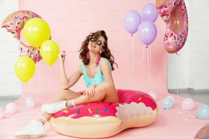Sommermode. Schöne Frau im modischen Badeanzug. Glückliches lächelndes weibliches Model mit sexy Körper in stilvoller Badebekleidung, die auf aufblasbarem Donut auf rosa Hintergrund sitzt. Hohe Auflösung.