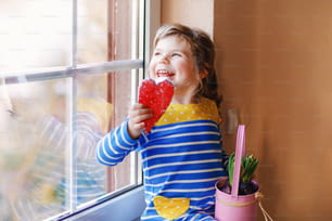 Little girl sitting by window with hyacinth flowers and hand-made heart. Happy child, indoors. Mother's day, valentine's day or birthday and spring concept
