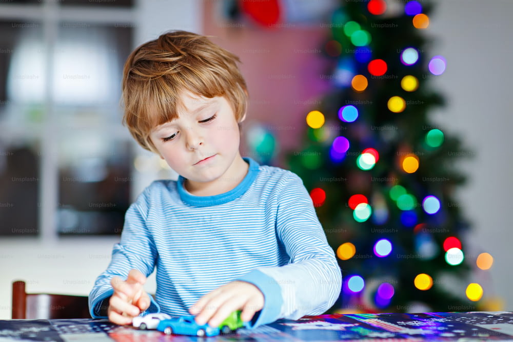 Pequeño niño rubio jugando con coches y juguetes en casa, en el interior. Lindo niño feliz divertido divirtiéndose con regalos. Luces de colores en el fondo. Concepto de tiempo de Navidad.