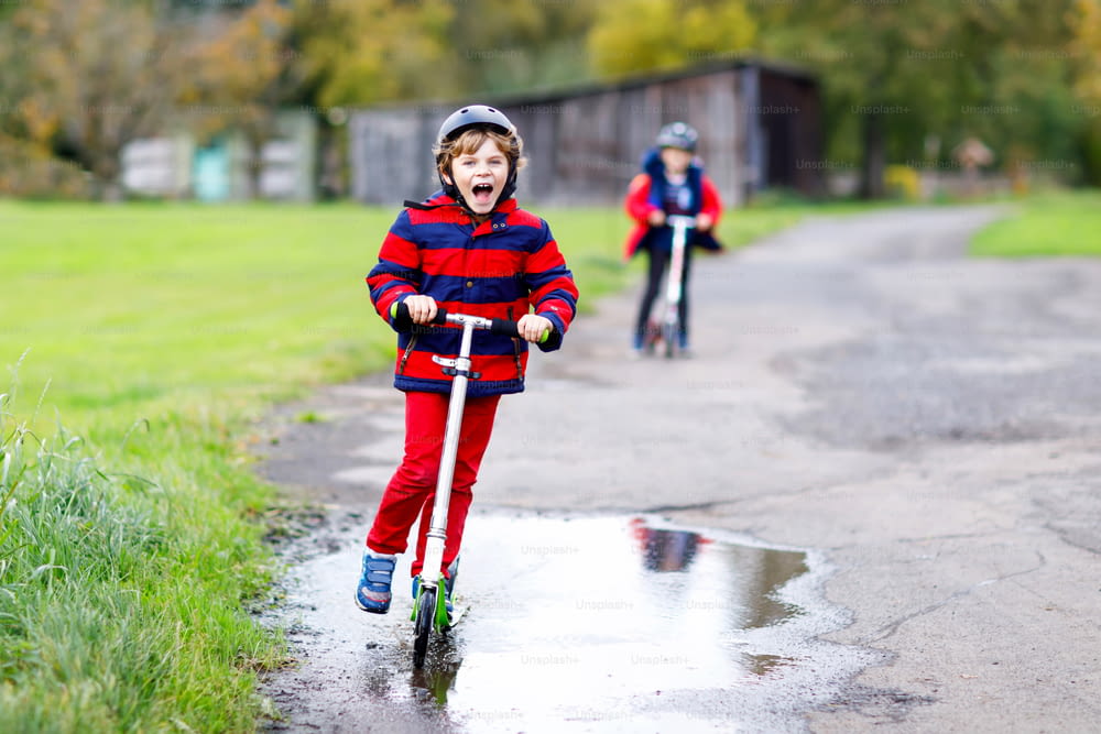 Dois meninos andando em patinetes empurram no caminho para a escola. Alunos de 7 anos dirigindo em meio a poças de chuva. Irmãos engraçados e melhores amigos jogando juntos