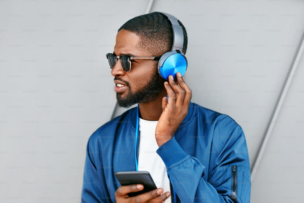 Escucha música. Hombre con auriculares y teléfono en ropa de moda. Guapo hombre negro escuchando música al aire libre. Alta resolución