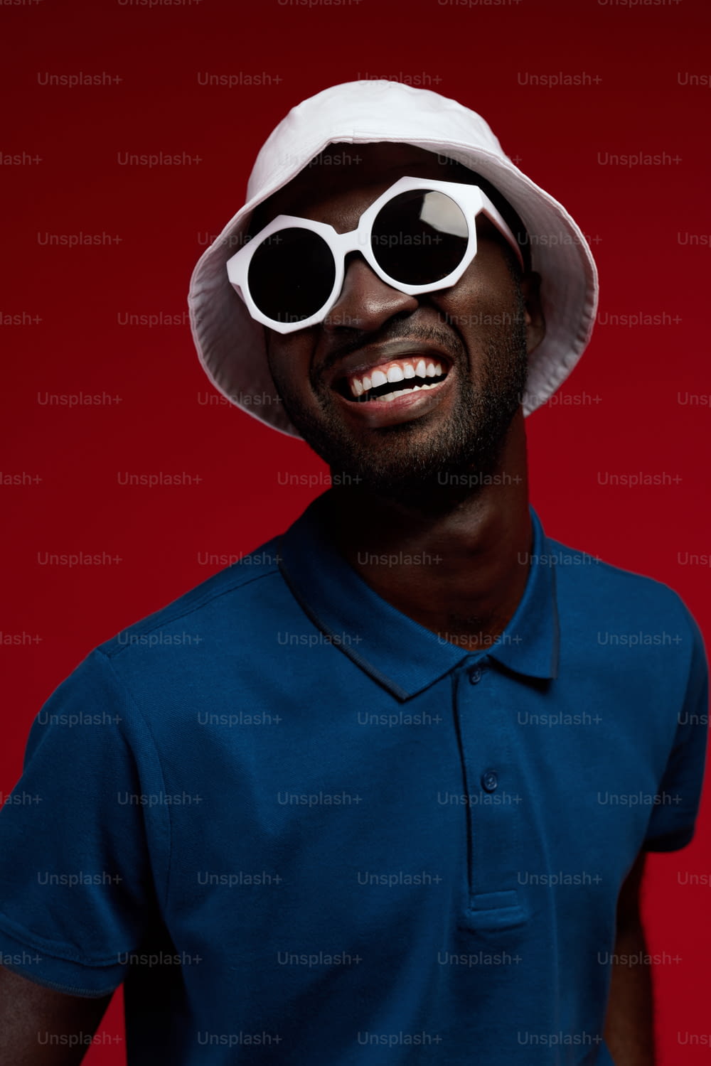 Modeporträt eines lächelnden schwarzen Mannes mit weißem Hut und Sonnenbrille auf rotem Hintergrund. Glückliches afroamerikanisches männliches Model in stylischem Sommeroutfit und Brille im Studio
