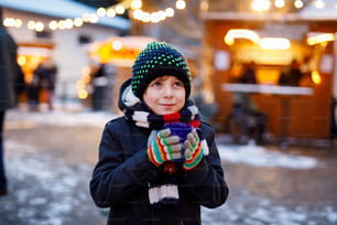 Little cute kid boy drinking hot children punch or chocolate on German Christmas market. Happy child on traditional family market in Germany, Laughing boy in colorful winter clothes.