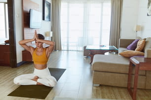 Meditación. Mujer en pose fácil practicando yoga en casa. Mujer joven sentada en el suelo y manteniendo las manos sobre la cabeza. Rutina deportiva para mantener la calma y la salud durante la cuarentena.