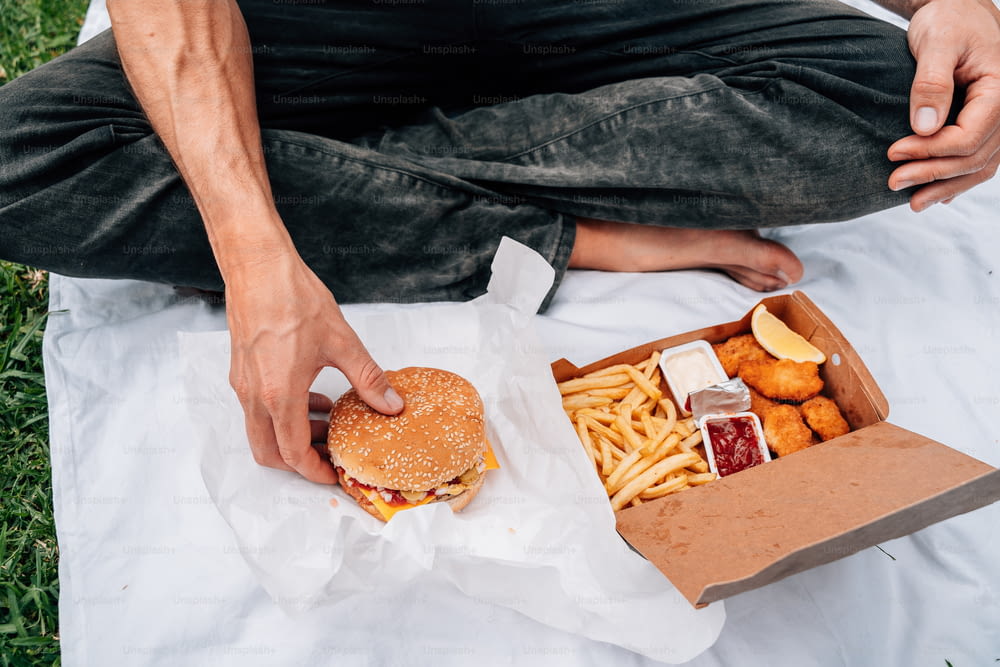 um homem sentado em um cobertor segurando um hambúrguer e batatas fritas