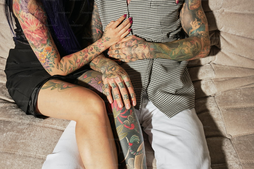 a man and woman sitting on a couch with tattoos on their arms