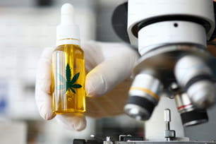 Focus on male hand in gloves holding glass bottle with cbd. Laboratory interior with microscope. Medicine and medical marijuana concept. Blurred background