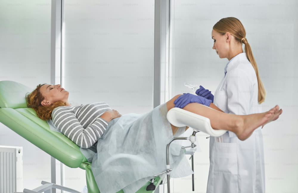 Vue latérale du portrait d’un jeune gynécologue en blouse blanche et gants stériles tenant un spéculum vaginal médical. Femme rousse aux yeux fermés allongée sur une chaise gynécologique