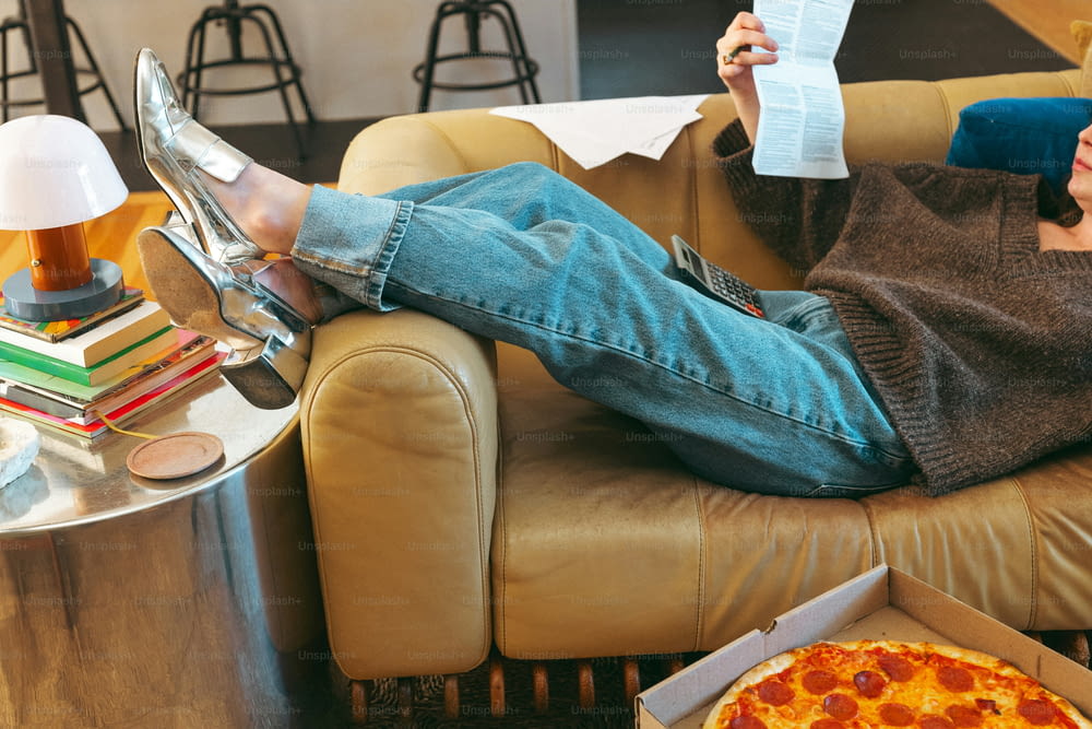 a woman sitting on a couch reading a paper and eating a pizza