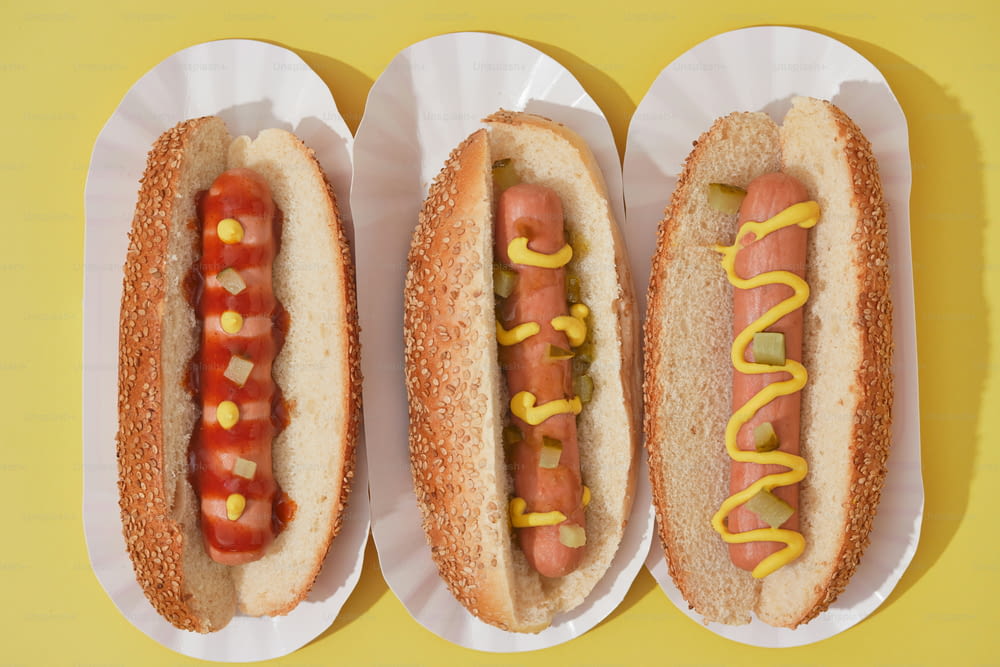 três cachorros-quentes com mostarda e ketchup em pratos de papel