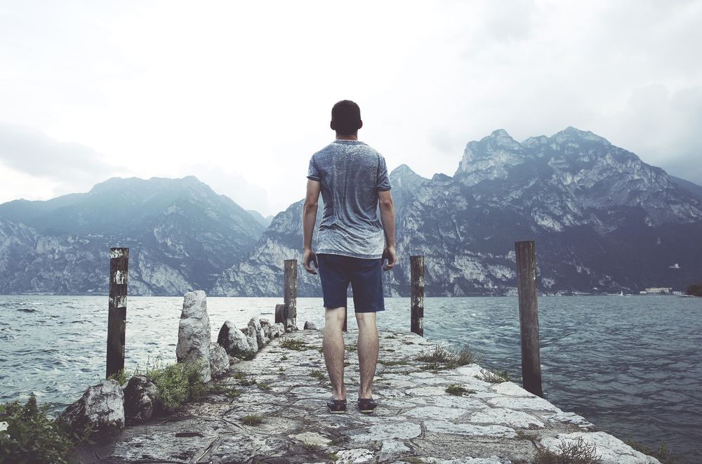 homme debout sur un quai en béton gris face à un plan d’eau et des montagnes pendant la journée
